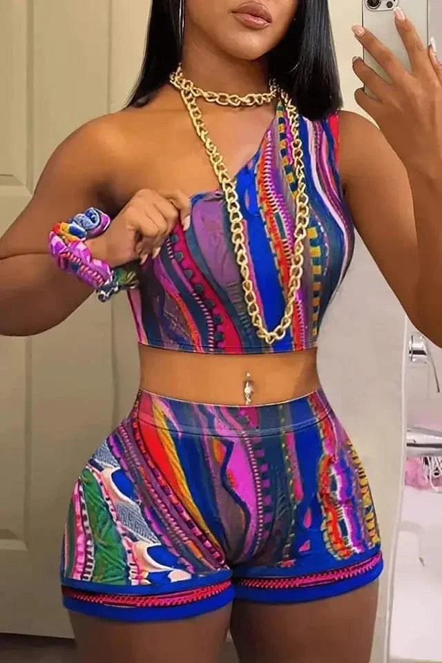 Woman wearing vibrant multicolor abstract print one-shoulder crop top and matching shorts with gold chain necklace and scrunchie